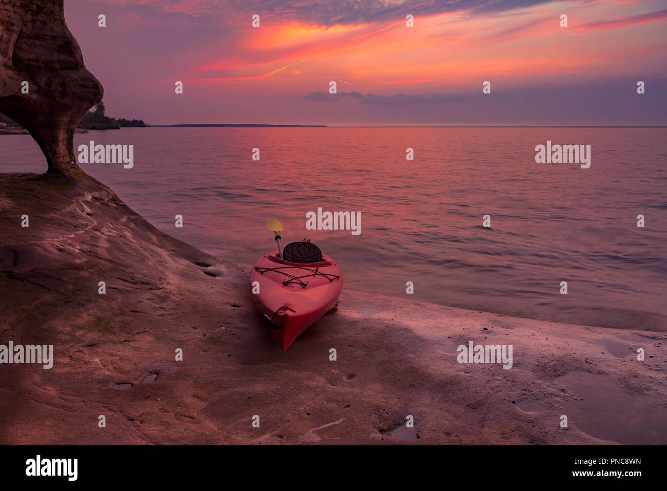 A red kayak sits on the rocky edge of a sea cave, under a vivid sunset on Lake Superior's shoreline in the Upper Peninsula of Michigan Stock Photo