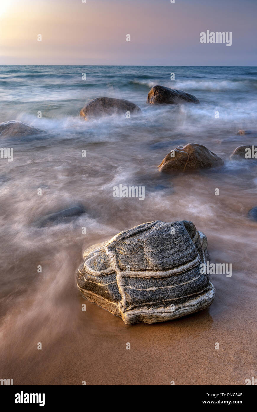 A unique rock and others glow under a Lake Superior sunset in the Upper Peninsula of Michigan. Waves ebb and flow in the golden beach light. Stock Photo