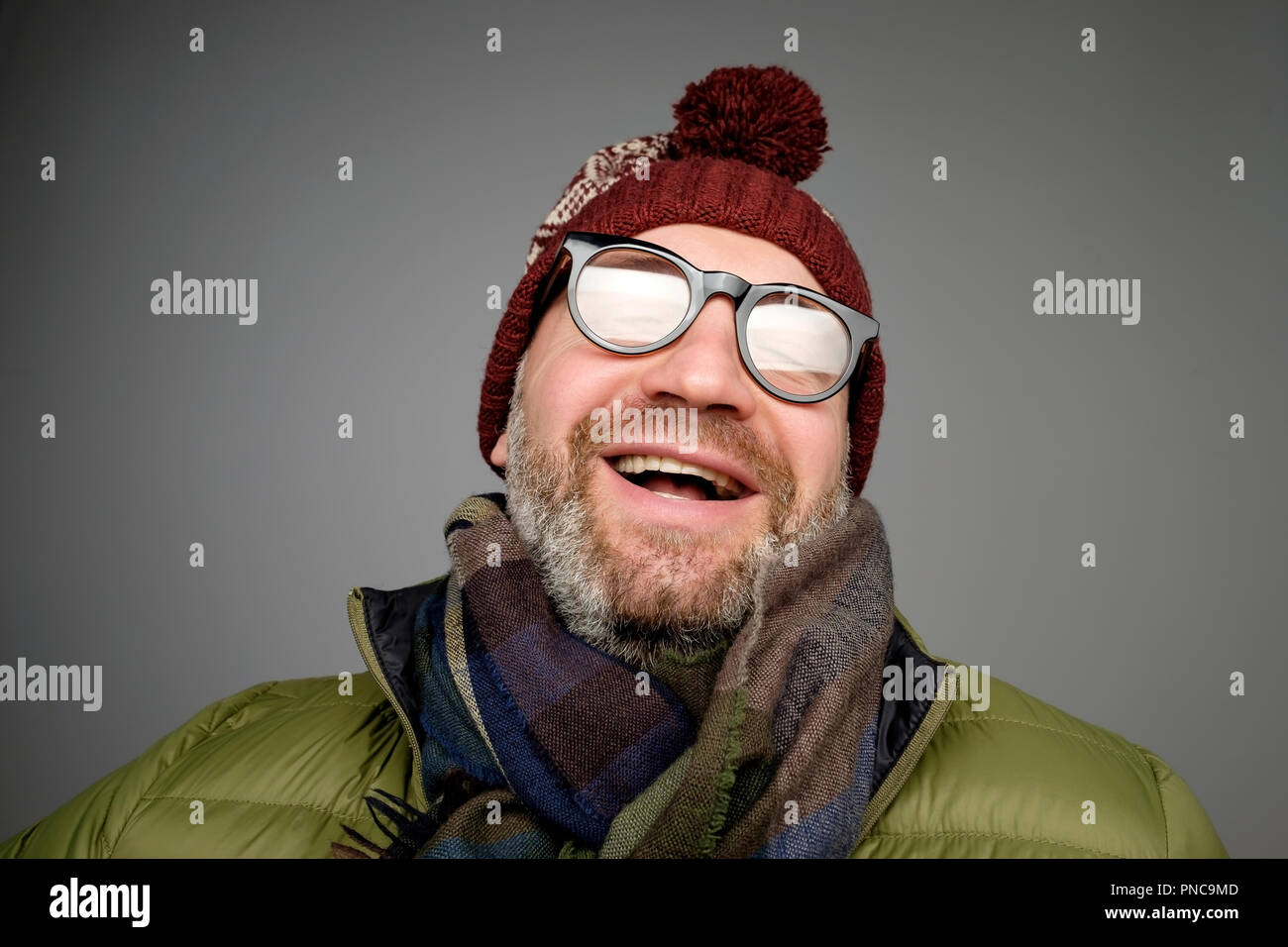 Portrait of mature hipster man in glasses and warm hat posing on gray background. Smiling bearded man wearing warm clothes for winter studio shot. Clo Stock Photo