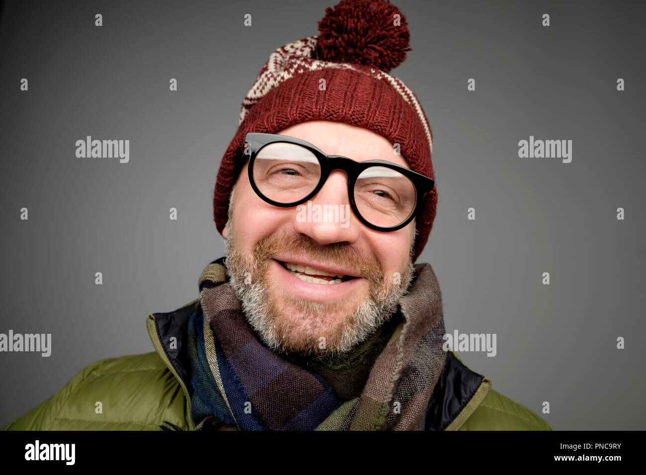 Portrait of mature hipster man in glasses and warm hat posing on gray background. Smiling bearded man wearing warm clothes for winter studio shot. Clo Stock Photo