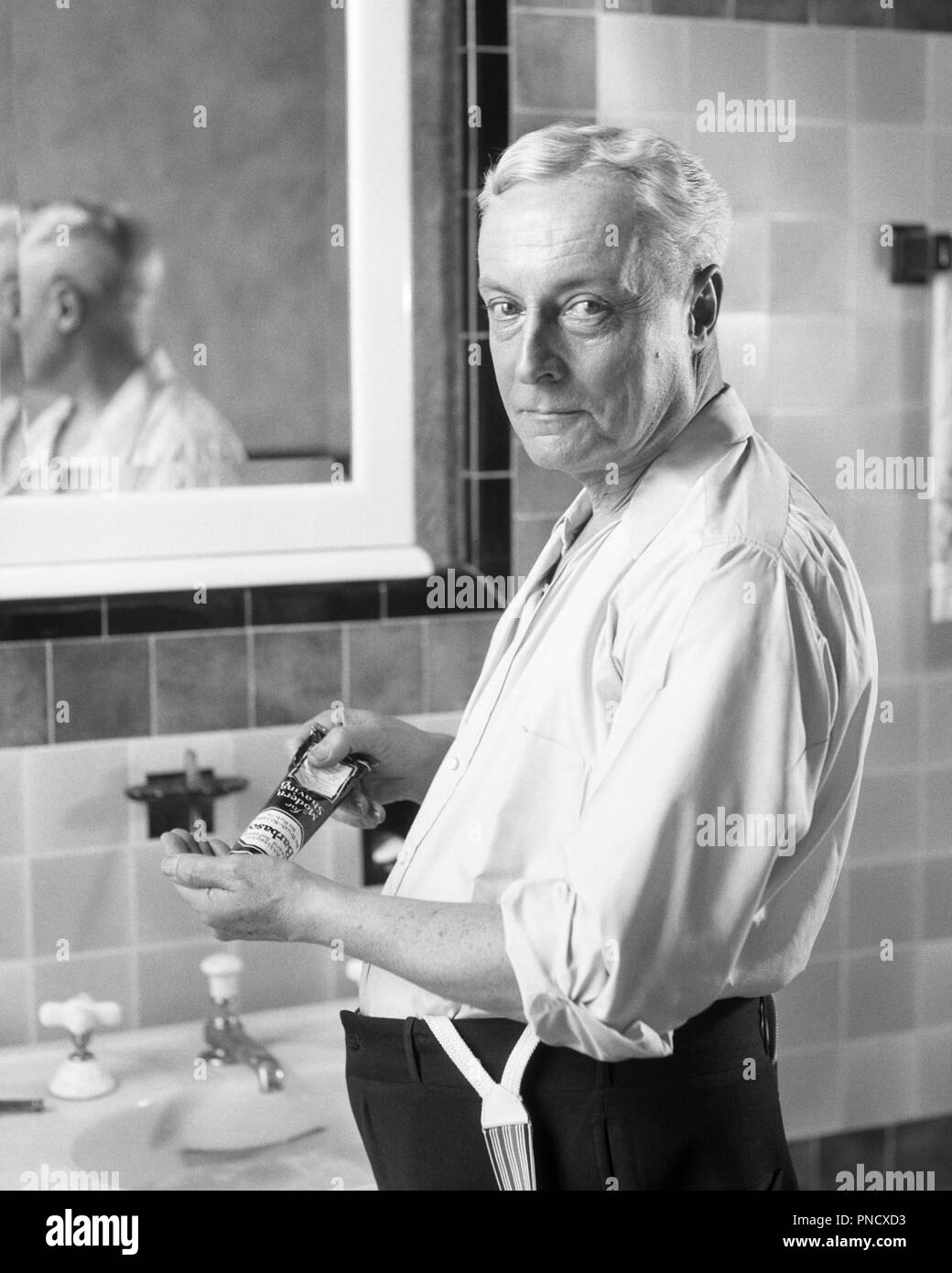 1920s SENIOR MAN STANDING IN SHIRT SLEEVES AT BATHROOM  SINK LOOKING AT CAMERA SQUEEZING GROOMING PRODUCT FROM TUBE INTO HAND   - b10305 HAR001 HARS EXPRESSIONS MIDDLE-AGED B&W MIDDLE-AGED MAN EYE CONTACT GROOMING BEFORE STARING OLDSTERS OLDSTER STRENGTH CHOICE KNOWLEDGE PRIDE AT IN INTO ELDERS SHIRT SLEEVES CONNECTION STARE STYLISH GRAY HAIR INTENSE PENETRATING SQUEEZING BLACK AND WHITE CAUCASIAN ETHNICITY HAR001 NEUTRAL OLD FASHIONED QUIZZICAL Stock Photo