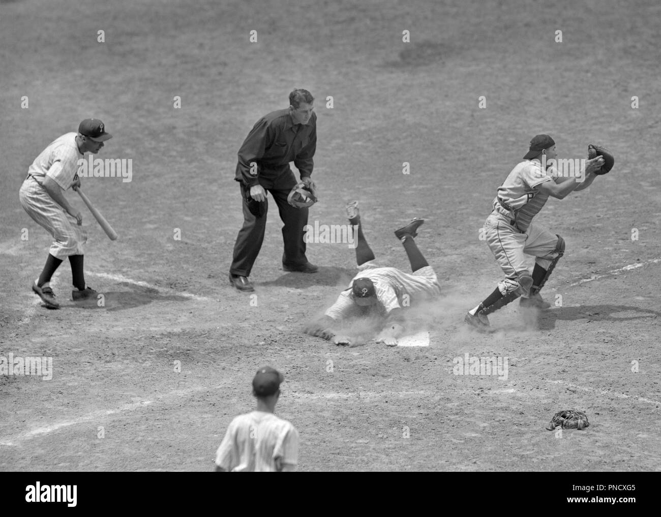 1950S 1960S baseball player safe at home plate cacther in front of plate umpire looking on next batter on deck - b11026 HAR001 HARS FULL-LENGTH PERSONS INSPIRATION MALES RISK NEXT B&W BATTER HIGH ANGLE UMPIRE CATCHER HOME PLATE AT IN OF ON OCCUPATIONS PROFESSIONAL SPORTS CONNECTION MOTION BLUR BALL GAME BALL SPORT MID-ADULT MID-ADULT MAN PHILLIES YOUNG ADULT MAN BASEBALL BAT BLACK AND WHITE CAUCASIAN ETHNICITY HAR001 OLD FASHIONED Stock Photo