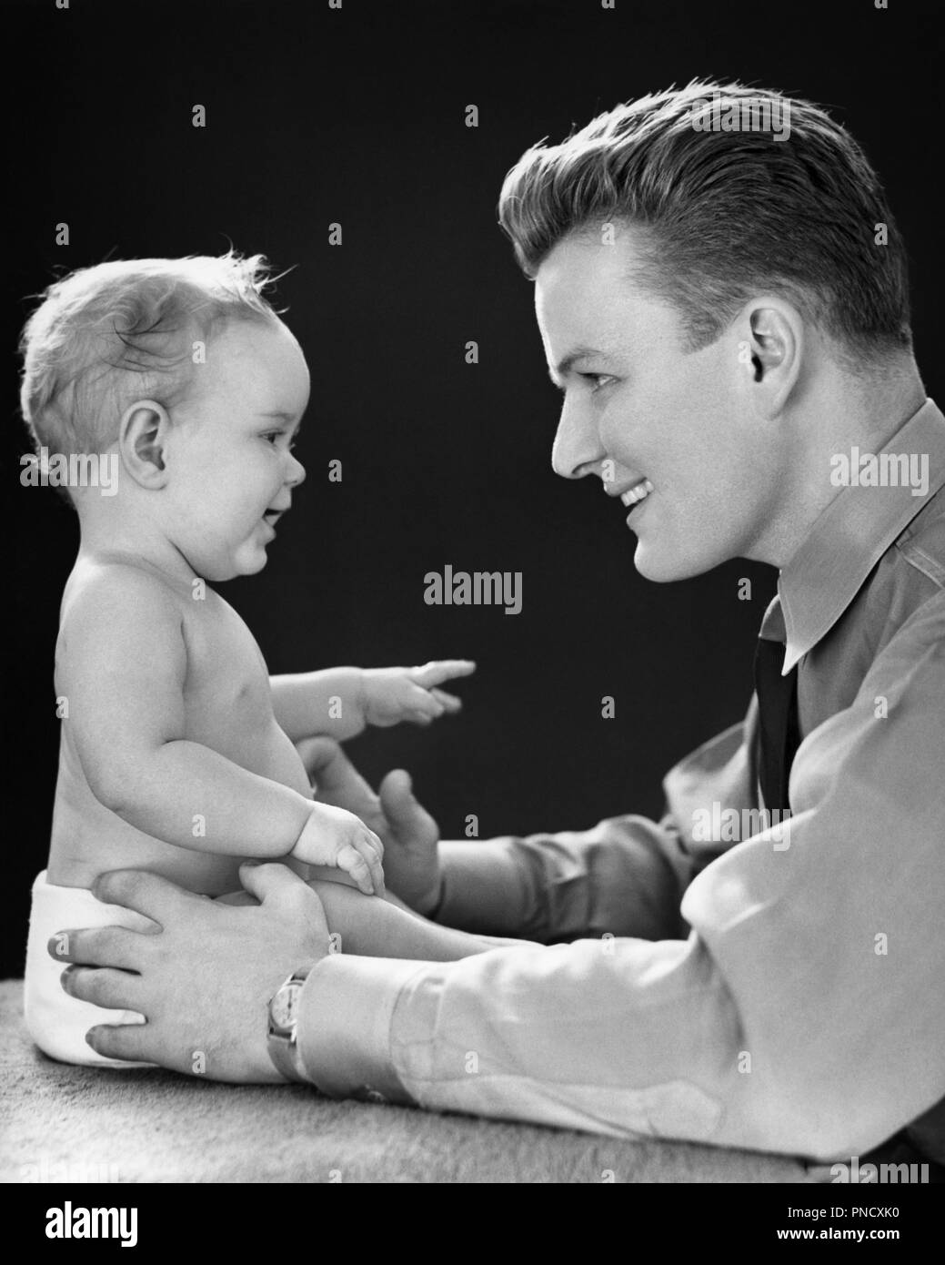 1940s SMILING YOUNG MAN FACE TO FACE EYE TO EYE LOOKING AT YEAR OLD BABY GIRL DAUGHTER SITTING UP  - b1168 HAR001 HARS LIFESTYLE CELEBRATION FEMALES HOME LIFE HALF-LENGTH DAUGHTERS PERSONS INSPIRATION CARING SPIRITUALITY CONFIDENCE FATHERS B&W HAPPINESS HEAD AND SHOULDERS CHEERFUL STRENGTH DADS EXCITEMENT POWERFUL PRIDE AT SMILES FACE TO FACE CONNECTION EYE TO EYE JOYFUL STYLISH GROWTH JUVENILES ONE YEAR OLD TOGETHERNESS YOUNG ADULT MAN BABY GIRL BLACK AND WHITE CAUCASIAN ETHNICITY HAR001 OLD FASHIONED SITTING UP Stock Photo