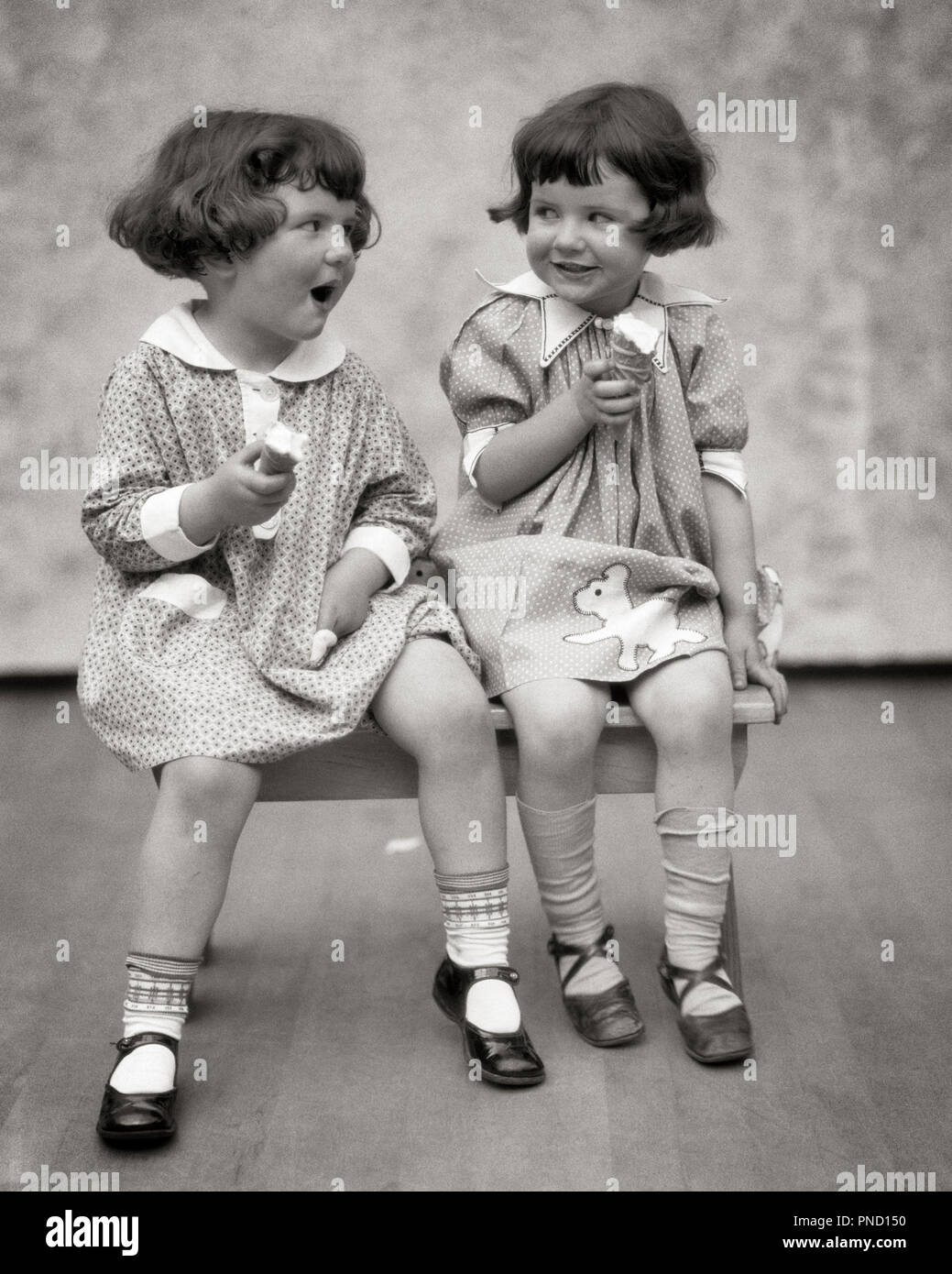 1920s TWO LITTLE GIRLS SITTING ON BENCH EATING ICE CREAM CONES AND TALKING  - f2181 HAR001 HARS COMMUNICATION LAUGH TWIN IDENTICAL DOUBLE PLEASED JOY FEMALES STUDIO SHOT HEALTHINESS HOME LIFE COPY SPACE FRIENDSHIP HALF-LENGTH MATCH SIBLINGS SISTERS EXPRESSIONS B&W BRUNETTE MATCHING SAME HAPPINESS CHEERFUL STYLES AND MARY JANE CONES SIBLING SMILES CONNECTION MARY JANES JOYFUL STYLISH LOOK-ALIKE DUPLICATE FASHIONS JUVENILES LOOK ALIKE TOGETHERNESS BLACK AND WHITE CAUCASIAN ETHNICITY CLONE HAR001 OLD FASHIONED PLAYMATES Stock Photo