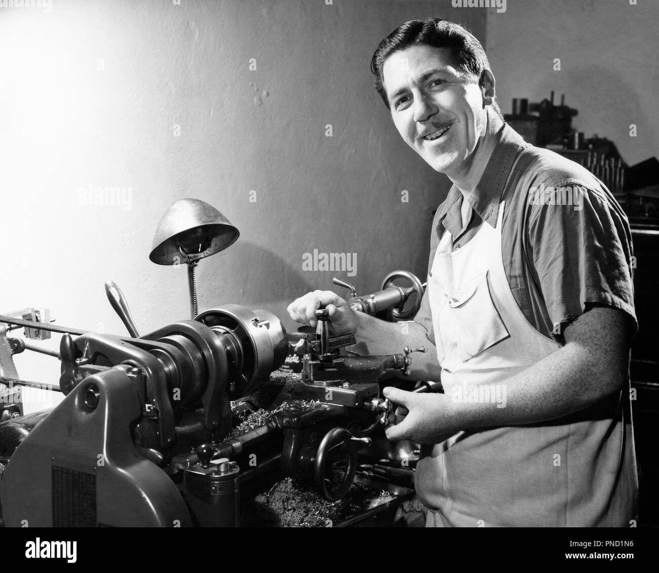 1950s PORTRAIT SMILING MAN FACTORY WORKER IN APRON WORKING WITH METAL ON LATHE LOOKING AT CAMERA  - i1223 DEB001 HARS PERSONS MALES CONFIDENCE EXPRESSIONS B&W EYE CONTACT SUCCESS MUSTACHE SKILL DECOR OCCUPATION HAPPINESS SKILLS CHEERFUL MACHINERY CUSTOMER SERVICE MUSTACHES KNOWLEDGE PRIDE AUTHORITY FACIAL HAIR OCCUPATIONS SMILES LATHE JOYFUL DEB001 MID-ADULT MID-ADULT MAN PRECISION BLACK AND WHITE CAUCASIAN ETHNICITY OLD FASHIONED Stock Photo