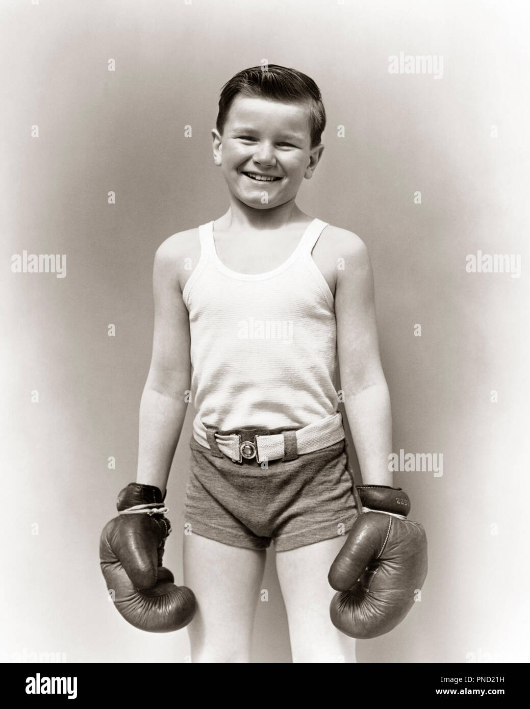 1930s SMILING BOY IN T-SHIRT AND GYM SHORTS STANDING LOOKING AT CAMERA  WEARING BOXING GLOVES READY FOR A FIGHT - j1977 HAR001 HARS FITNESS SHORTS  JUVENILE STYLE FEAR HEALTHY BALANCE SAFETY COMPETITION
