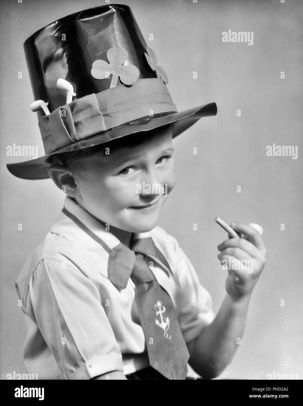 1930s IMPISH YOUNG IRISH LAD SMILING LOOKING AT CAMERA WEARING TOP HAT DECORATED WITH SHAMROCKS HOLDING TRADITIONAL CLAY PIPE  - j6719 HAR001 HARS HALF-LENGTH INSPIRATION IRELAND TRADITIONAL CHARACTER MALES CONFIDENCE DECORATED EXPRESSIONS B&W EYE CONTACT SHAMROCK HAPPINESS HEAD AND SHOULDERS CHEERFUL CLAY PIPES STRENGTH PRIDE MISCHIEVOUS SMILES CONNECTION CONCEPTUAL JOYFUL STYLISH TYPE IMPISH LITTLE PEOPLE JOKING JUVENILES LEPRECHAUN STEREOTYPE BLACK AND WHITE CAUCASIAN ETHNICITY GAELIC HAR001 ICONIC INNOCENT OLD FASHIONED SHAMROCKS Stock Photo