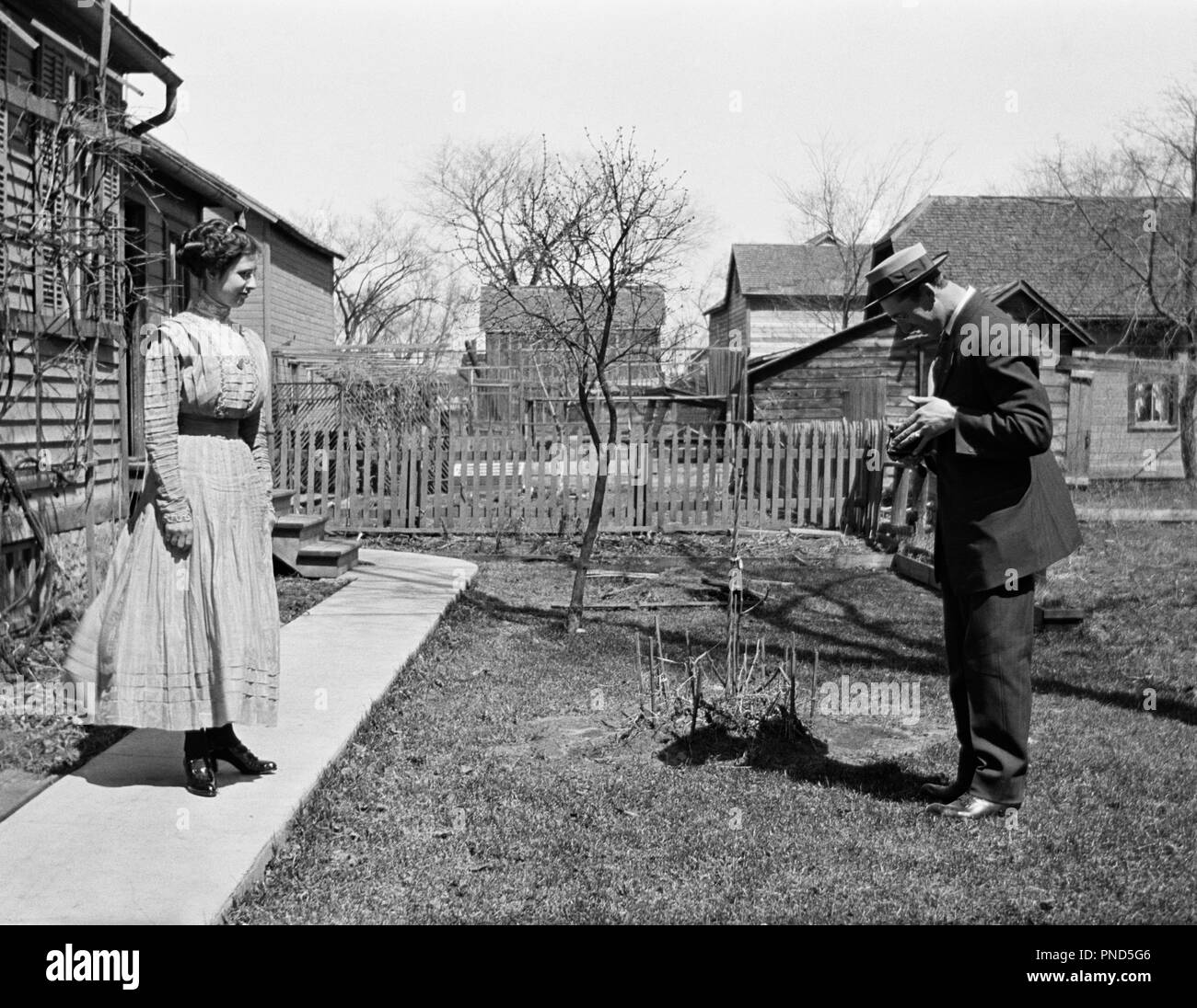 1900s 1910s TURN OF THE 20TH CENTURY MAN TAKING PICTURE OF WOMAN STANDING IN FRONT OF HOUSE  - o3633 LEF001 HARS YOUNG ADULT PHOTOS TURN PHOTOGRAPHS LIFESTYLE FEMALES MARRIED RURAL SPOUSE HUSBANDS GROWNUP HOME LIFE HORIZONTAL COPY SPACE FULL-LENGTH LADIES PERSONS CAMERAS MALES CAMERAMAN B&W MEN AND WOMEN PARTNER PICTURES PHOTOGRAPHERS LEISURE TURN OF THE 20TH CENTURY GENTLEMAN AND GENTLEMEN TURN OF THE CENTURY OF THE PHOTOGRAPHIC MANLY PHOTOGRAPHY CAMERAMEN PARTNERS TOGETHERNESS WIVES YOUNG ADULT MAN YOUNG ADULT WOMAN BLACK AND WHITE CAUCASIAN ETHNICITY OLD FASHIONED Stock Photo