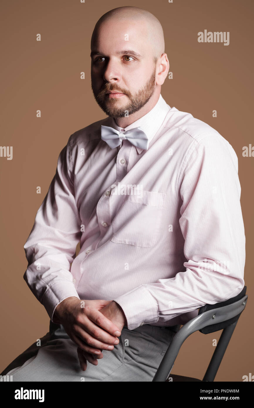 Profile side view portrait of handsome bearded bald man in light pink shirt and white bow, sitting on chair and looking away with thoughtful serious f Stock Photo