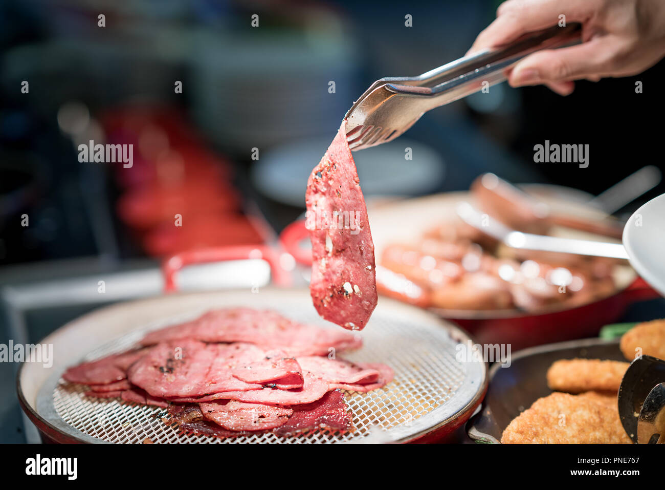 Human hand holding tongs over plate of sliced smoked meat in bar at restaurant in hotel. Breakfast appetizer in plate. Stock Photo