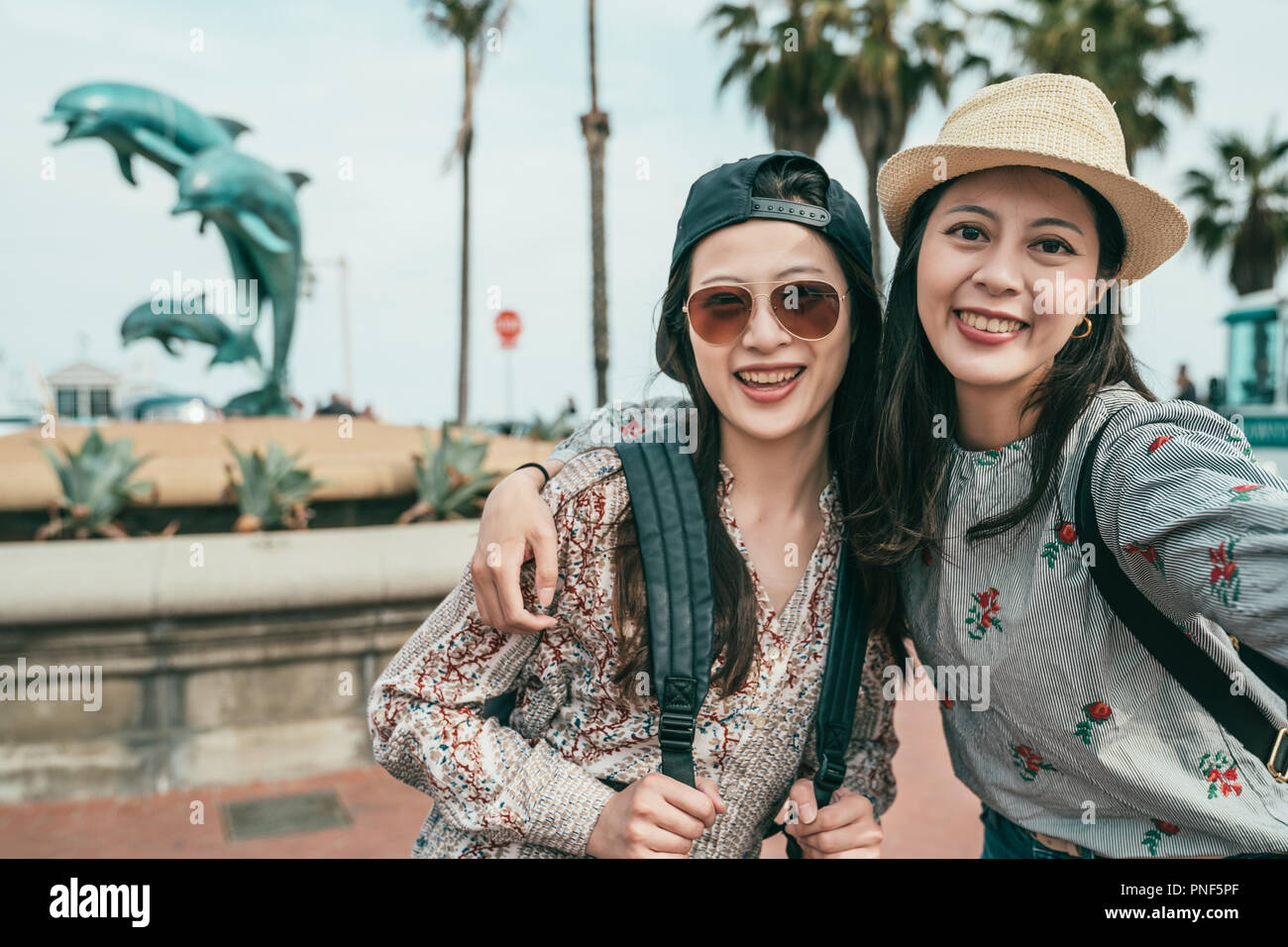 two women smiling and taking selfies happily in front of a dolphin fountain in a lovely plaza. Stock Photo