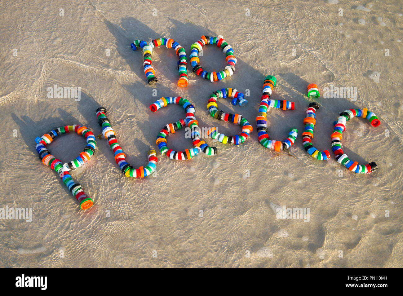 Colorful bottle caps spell out 'no plastic' on a sandy beach as a wave laps around. A reminder for people to take action on pollution Stock Photo