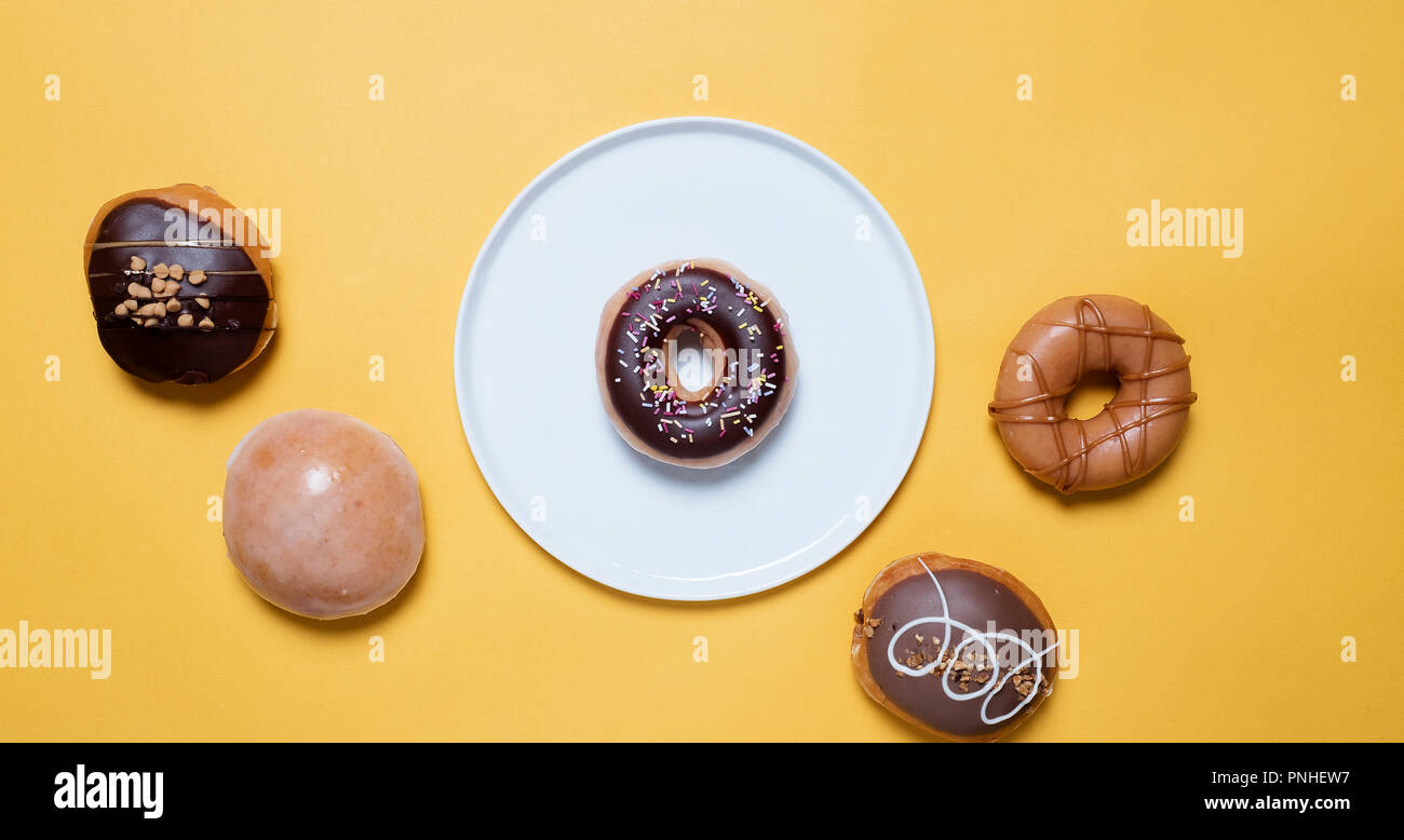 Classic chocolate donuts with sprinkles on a white plate on a yellow background surrounded by various other decorated donuts Stock Photo