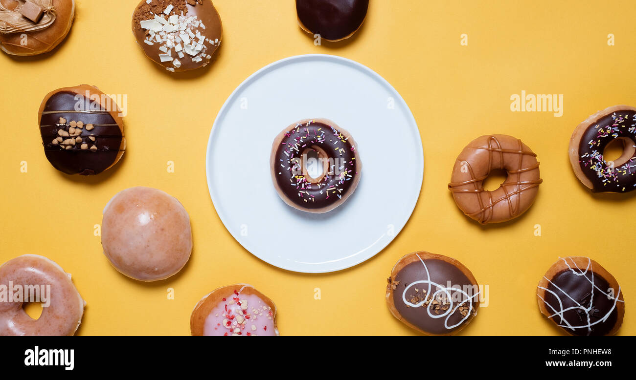 Classic chocolate ring donuts with sprinkles on a white plate on a yellow background surrounded by various other decorated donuts with space for copy. Stock Photo