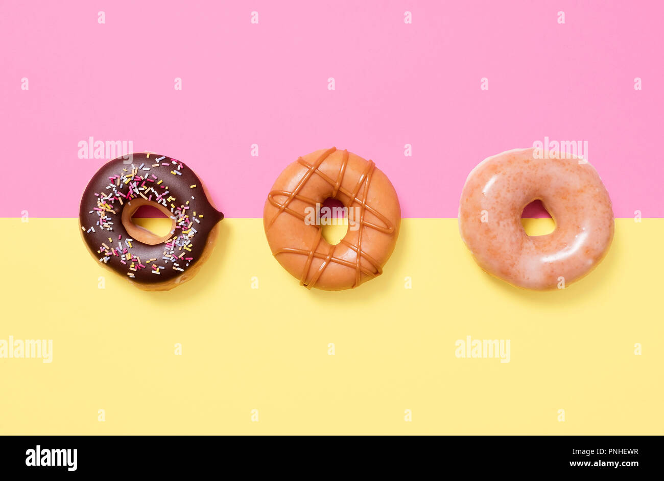 Three donuts in a row on a yellow and pink split background with chocolate sprinkles, plain, and swirled caramel Stock Photo