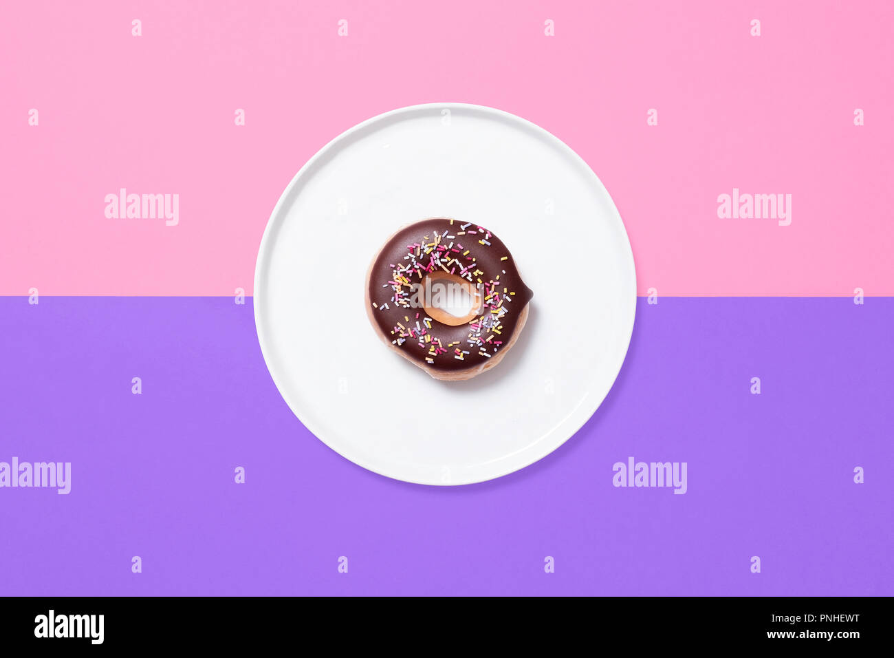 Classic ring donut with chocolate glaze frosting and sprinkles on a white plate on a split coloured pastel background of pink and purple. Stock Photo