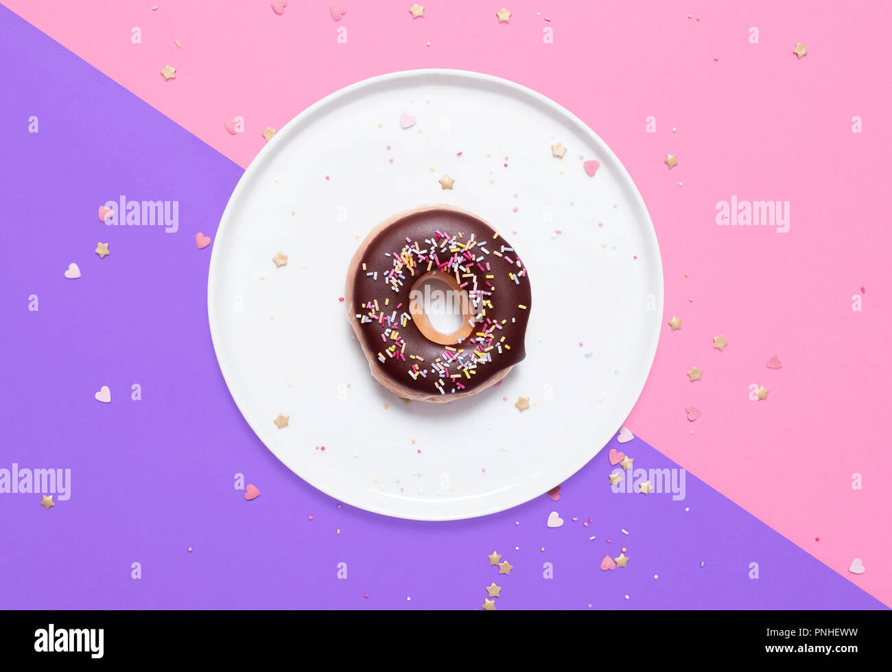 Classic donut with chocolate glaze frosting and sprinkles on a white plate on a split pink and purple pastel background with hearts scattered Stock Photo