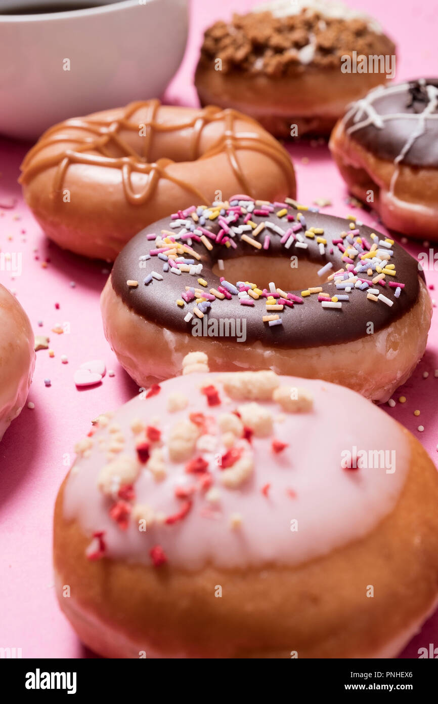 Assorted donuts on a pastel pink background with cup of coffee in the background.  Includes chocolate ring with sprinkles as well as various others Stock Photo