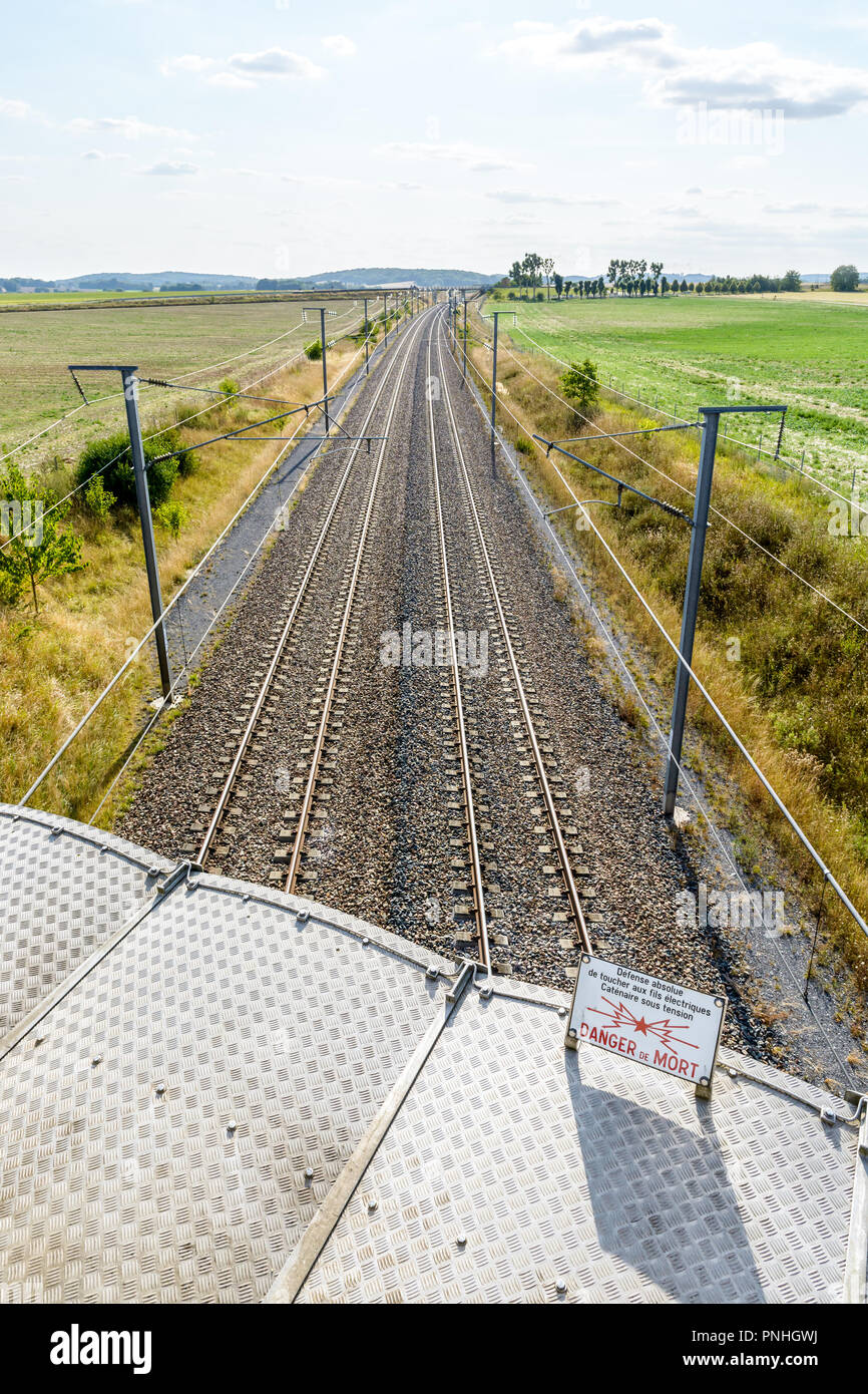 A french high speed railway with overhead line equipment (posts, catenaries and power lines) to supply trains and a danger sign in the foreground. Stock Photo