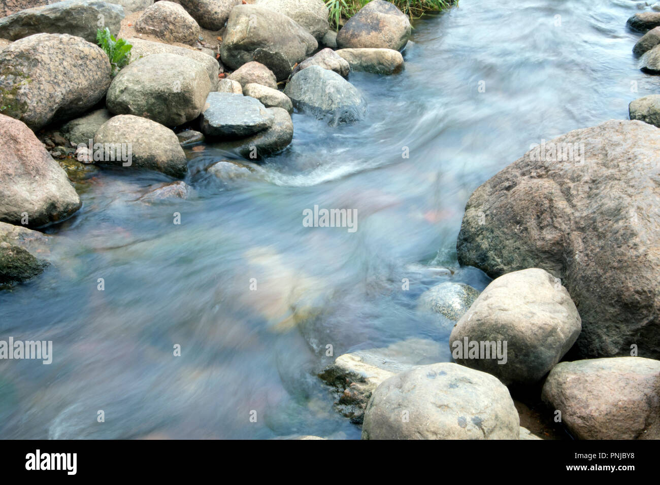 Small creek with stony bottom, rapid stream running between boulders, smoothed water surface Stock Photo