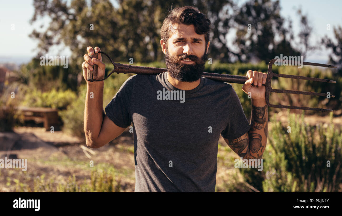 Beard man with pitchfork standing in the farm looking at camera. Male farmer with pitch fork standing outdoors on his on allotment. Stock Photo