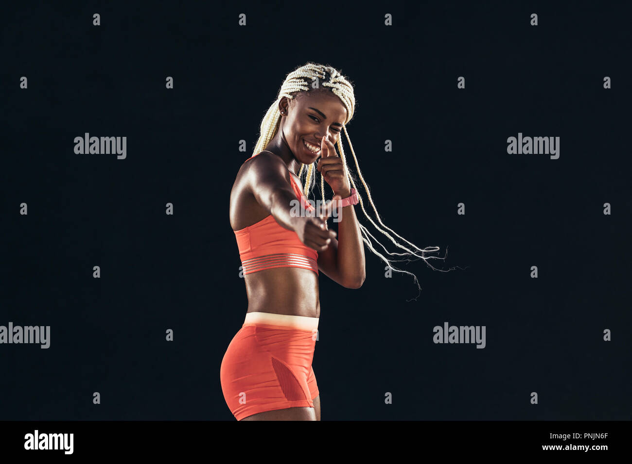 Smiling woman athlete in fitness attire making a pointing hand gesture winking an eye. Portrait of a female sprinter in happy mood during training. Stock Photo