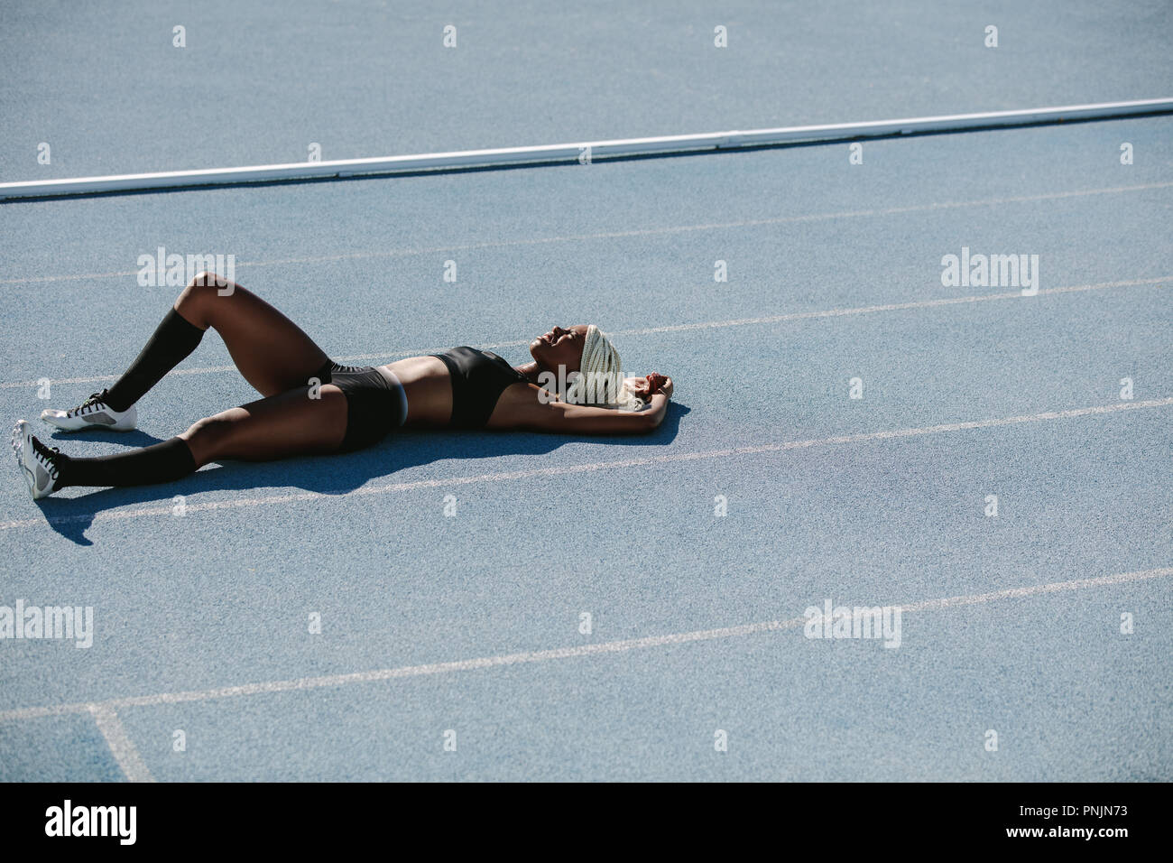 Woman athlete lying on the running track relaxing after training. Female sprinter resting after workout lying on track on a sunny day. Stock Photo