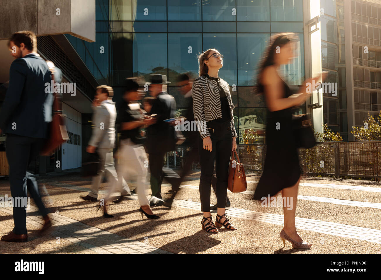 Businesswoman holding her hand bag standing still on a busy street with people walking past her using mobile phones. Woman standing amidst a busy offi Stock Photo