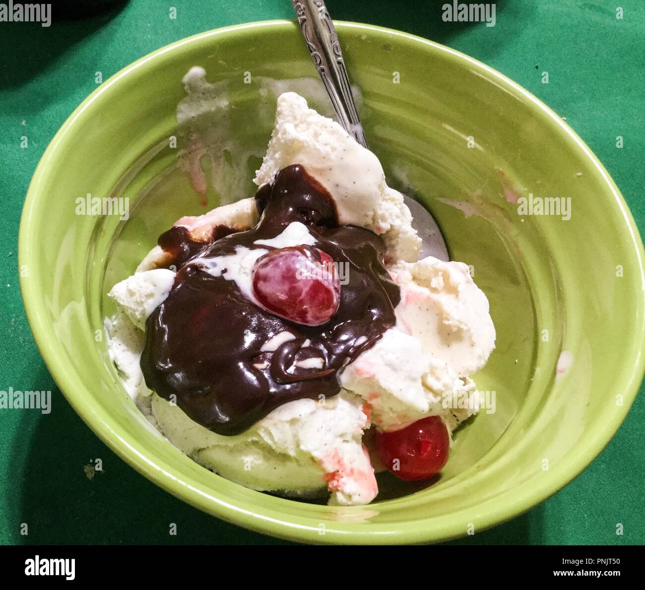 Ice cream with banana, cherries and hot fudge sauce in light green bowel with spoon on dark green background. Stock Photo