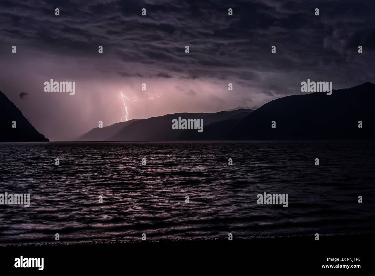 Lightning in dark blue clouds during a thunderstorm over a lake in the mountains Stock Photo
