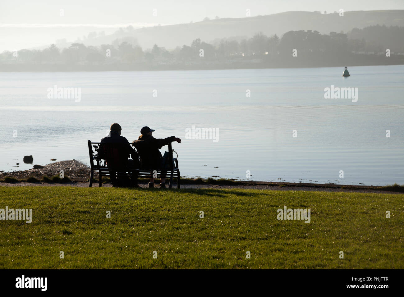 One man and one woman sitting on a bench over looking the Gareloch, Scotland with mist in the background in autumn. Stock Photo