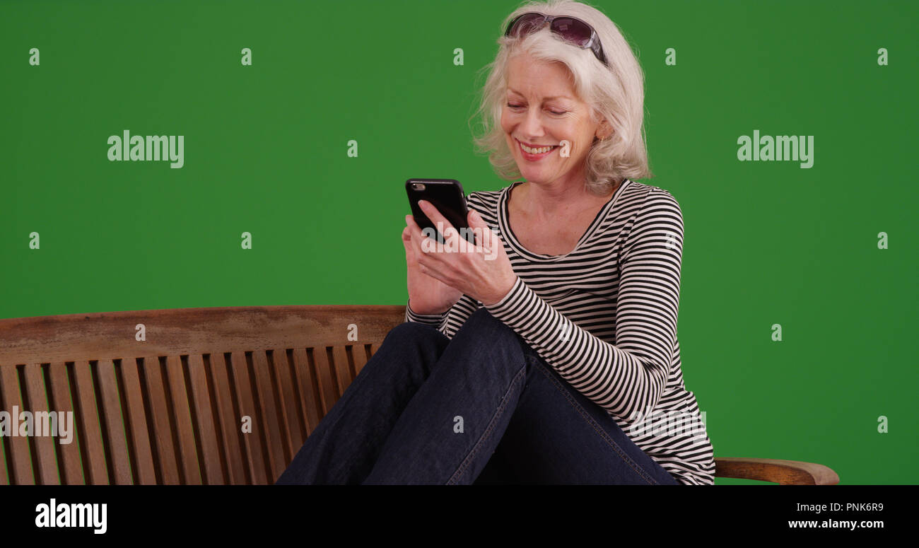 Smiling old white female using mobile device on park bench on green screen Stock Photo