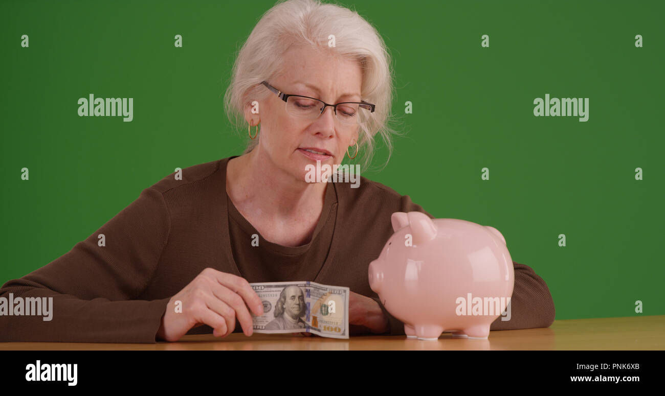 Elderly woman about to put 100 dollar bill into piggy bank on green screen Stock Photo