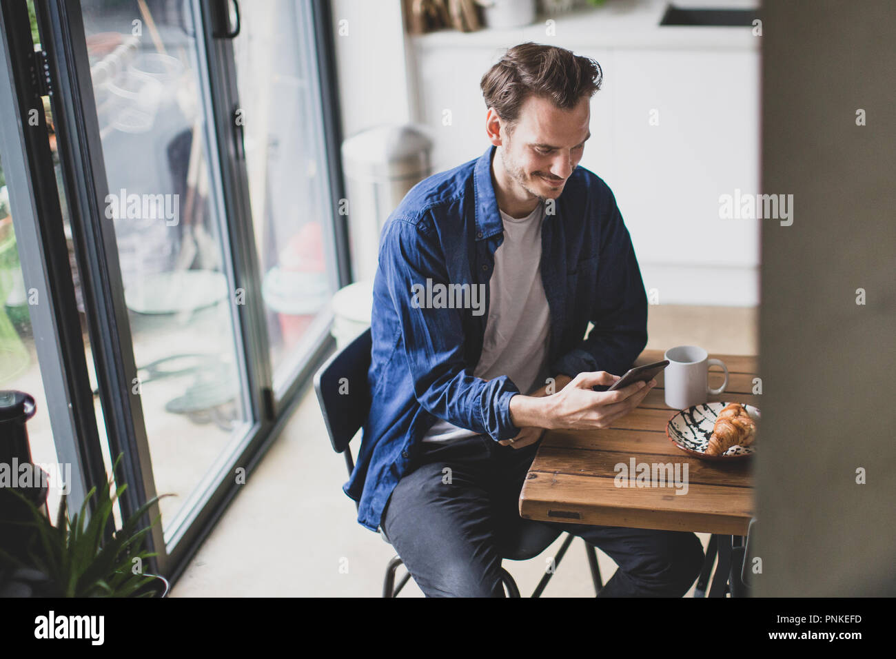 High angle shot of adult male checking smartphone in kitchen with mug of coffee Stock Photo