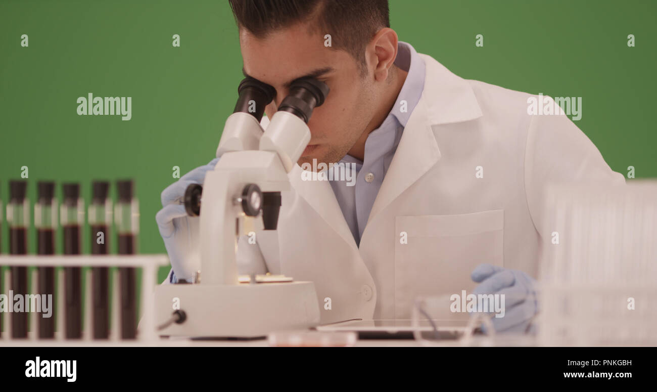 Millennial Hispanic medical scientist looking through microscope on green screen Stock Photo