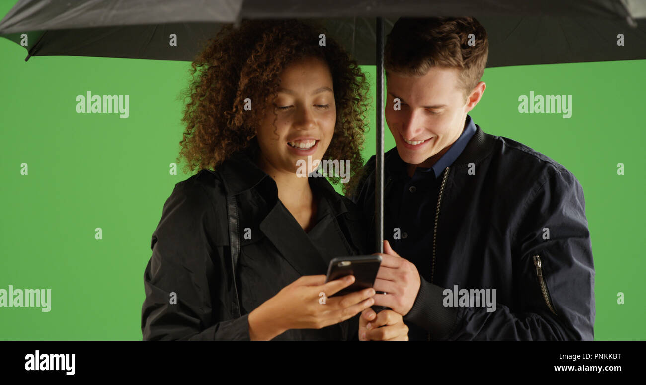 Young couple sharing umbrella in the rain using phone on green screen Stock Photo