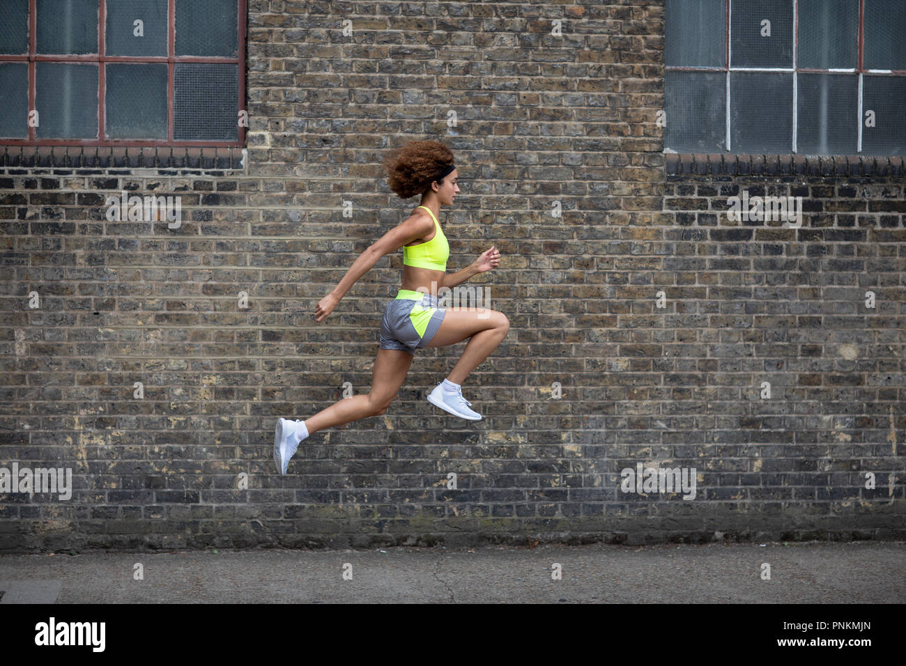 Young adult female leaping in the air with brick wall backdrop Stock Photo