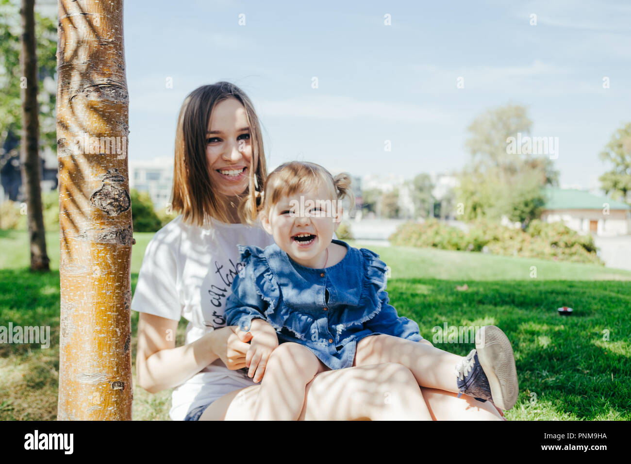 Portrait of a young mother and her 3 years old daughter sitting under the  tree in a city park on a warm sunny day Stock Photo - Alamy