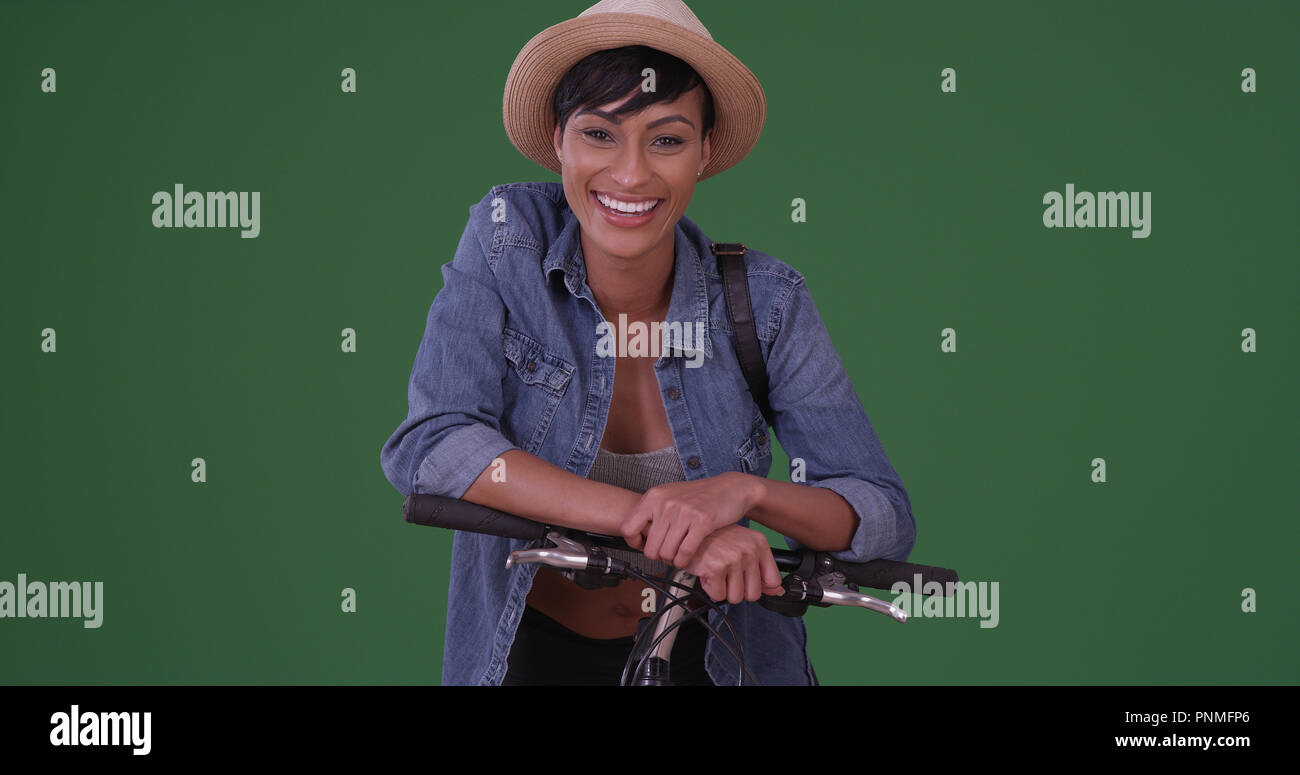 Young black hipster wearing sunhat resting on bike and smiling on green screen Stock Photo