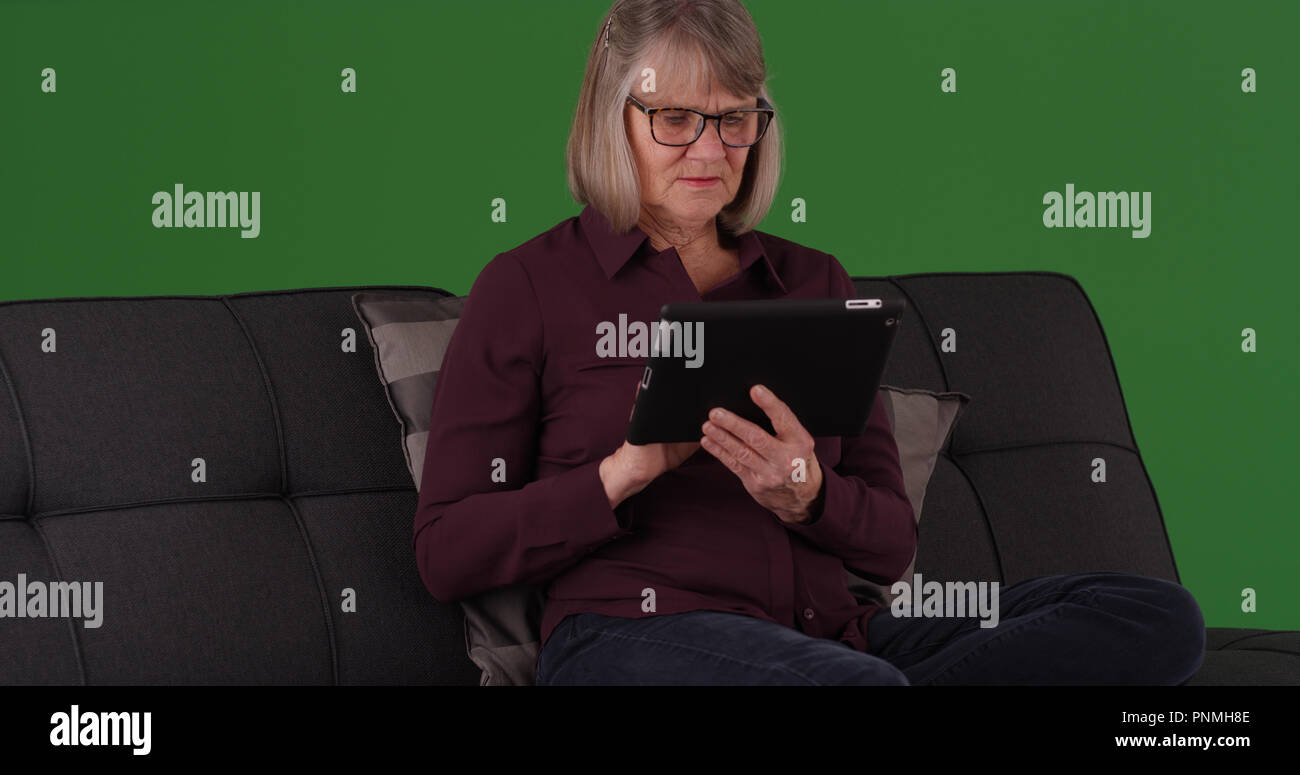 Modern elderly female sitting on couch using tablet device on green screen Stock Photo