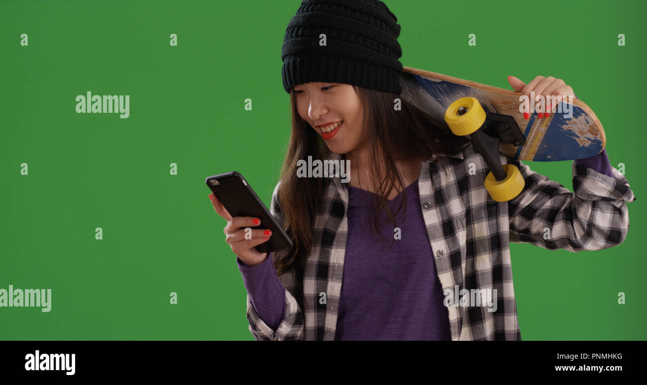 Asian skater girl carrying skateboard on shoulder using phone on green