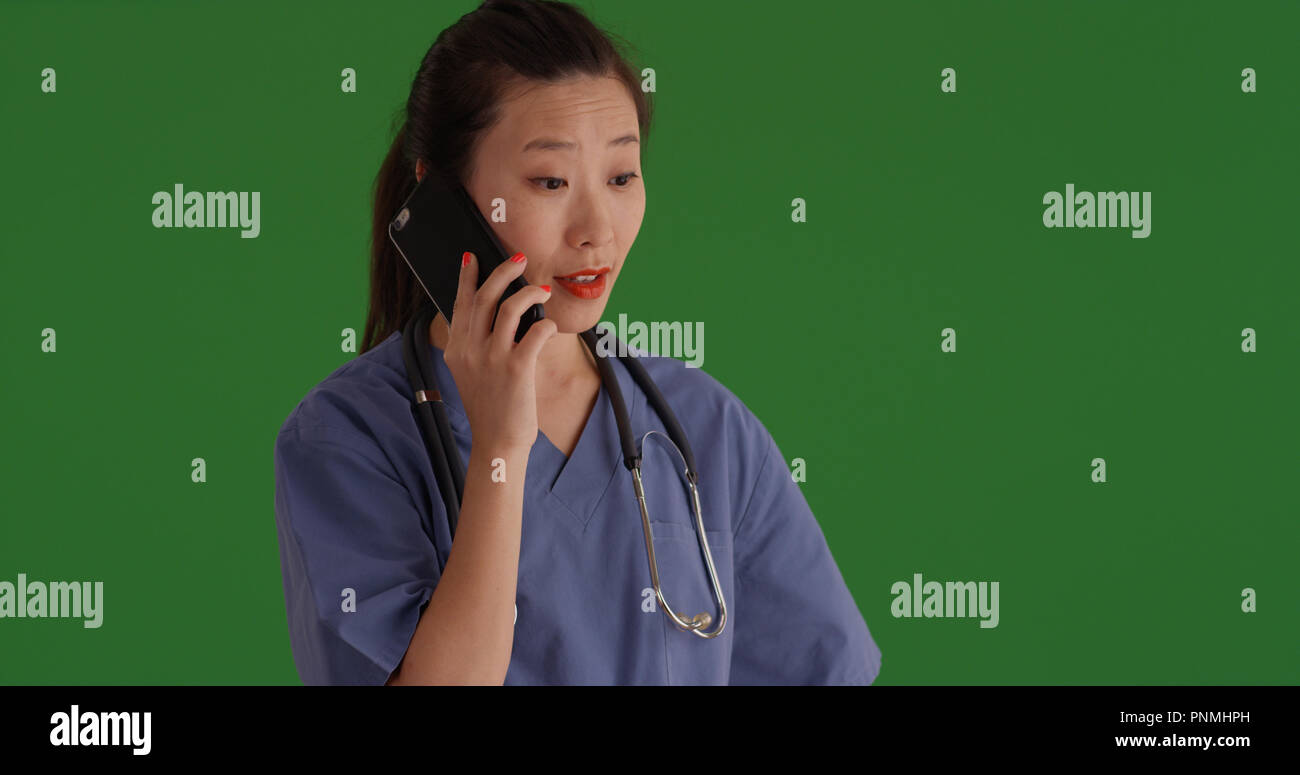 Asian female doctor in scrubs talking on smartphone on green screen ...