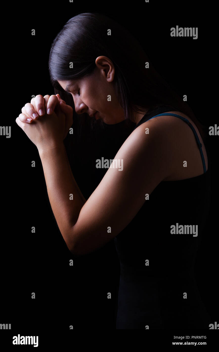 Faithful woman praying, hands folded in worship to god with head down and eyes closed in religious fervor, on a black background. Concept for religion Stock Photo