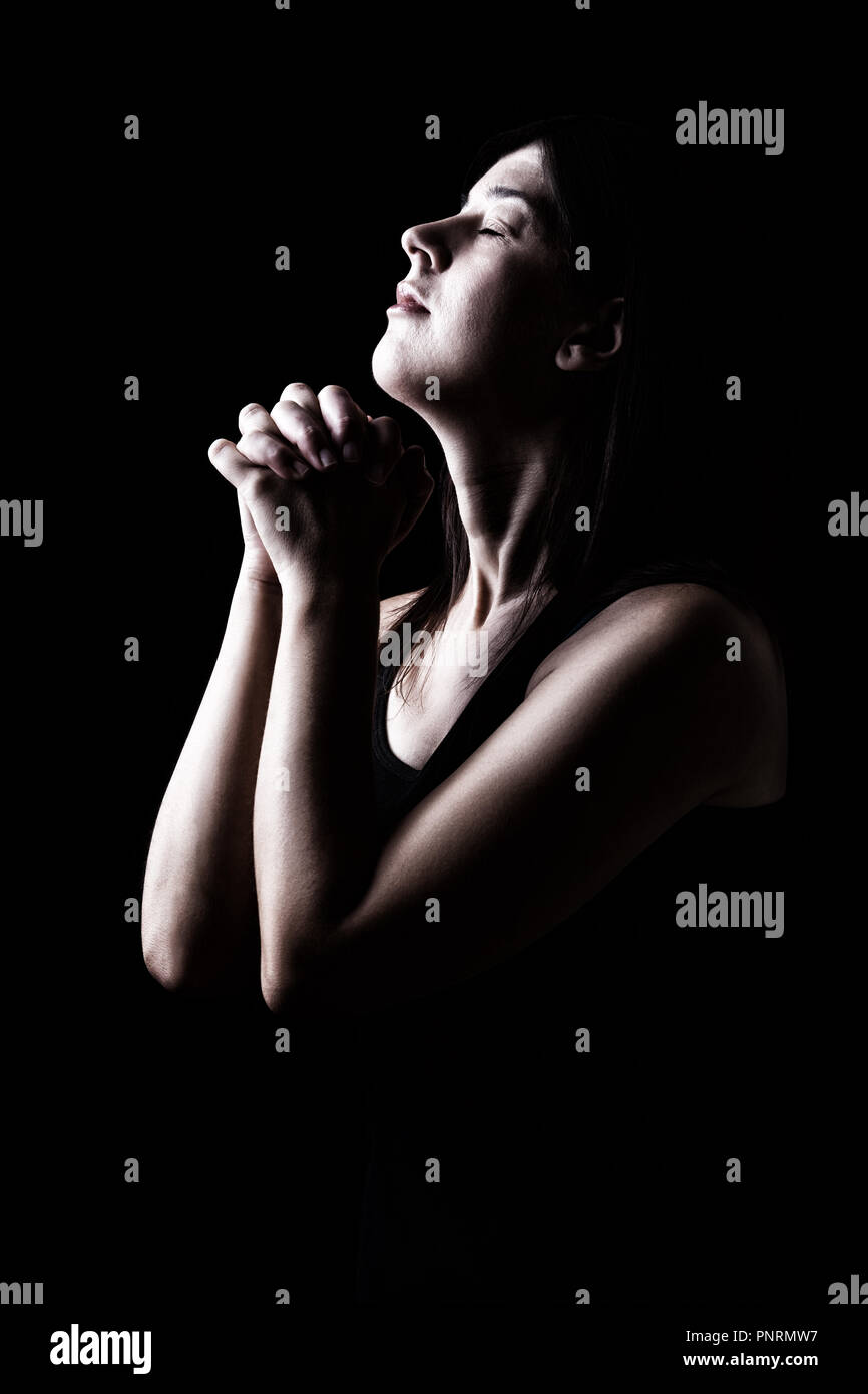 Faithful woman praying, hands folded in worship to god with head up and closed eyes in religious fervor, on a black background. Concept for religion,  Stock Photo
