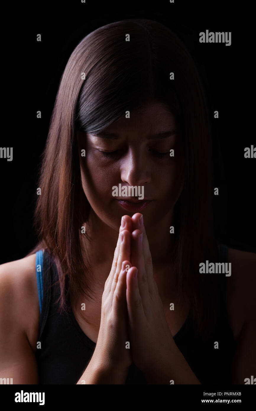 Faithful athletic woman praying, with hands folded in worship to god, head down and eyes closed in religious fervor, on black background. Concept for  Stock Photo