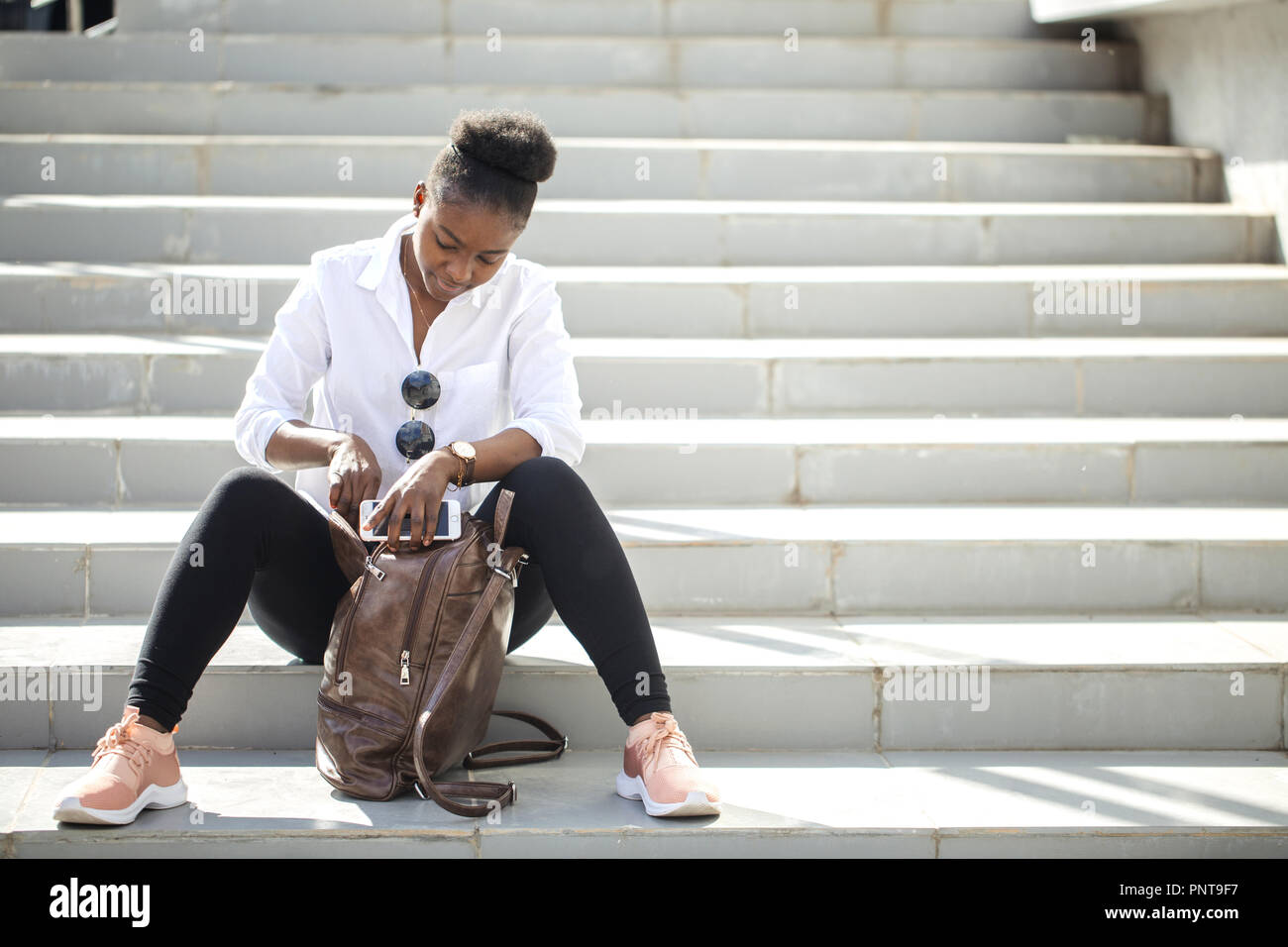 African woman using smart phone while sitting on white stairs outdoors. Stock Photo