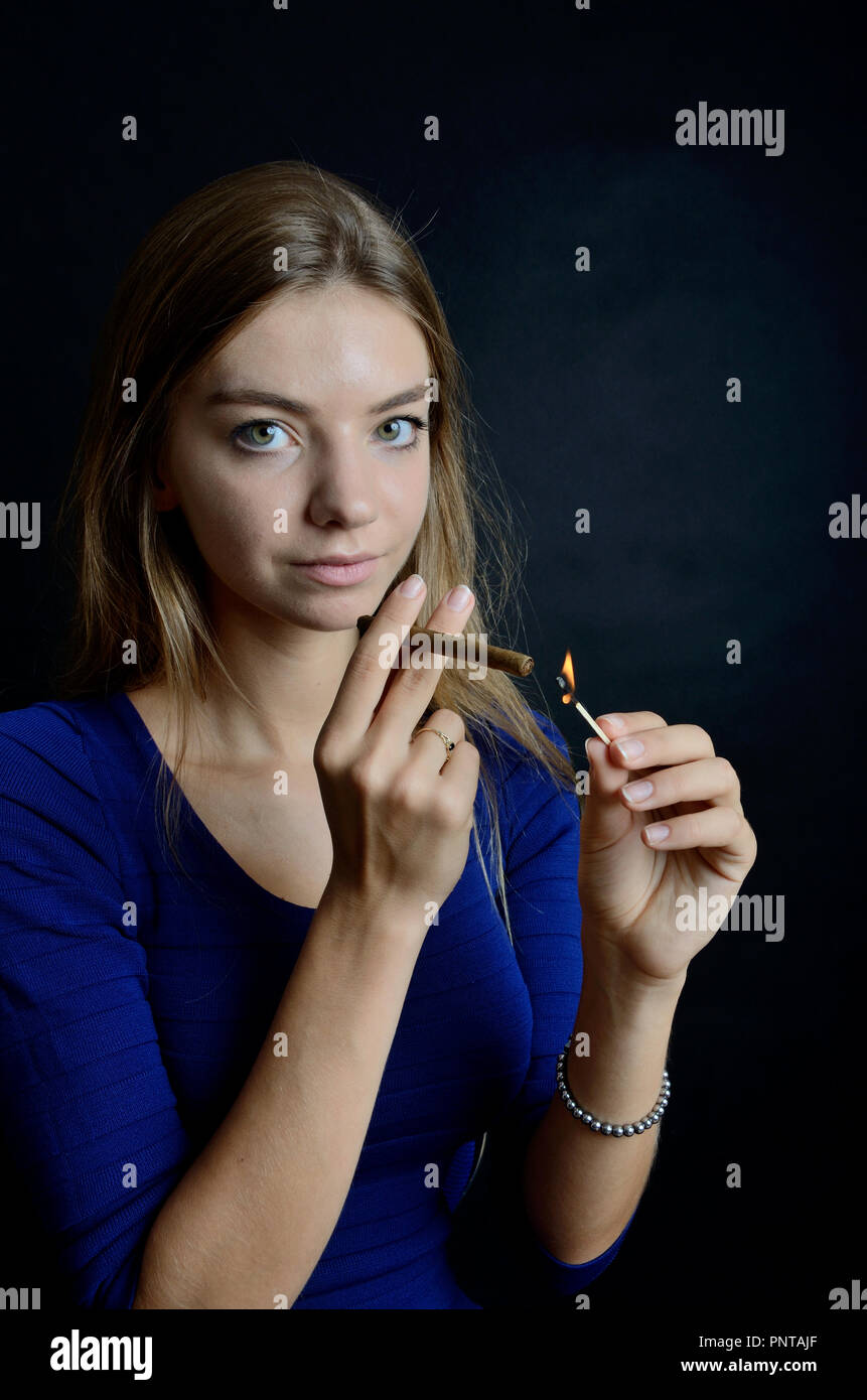 Young female lighting up cigarillo with matches. Woman in blue dress with black background. Stock Photo