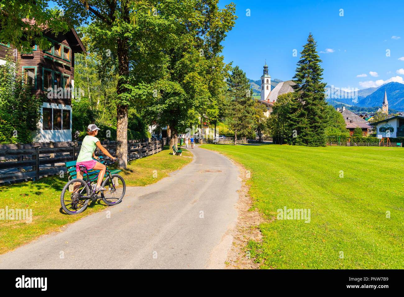 KITZBUHEL TOWN, AUSTRIA - JUL 30, 2018: Woman on bike on path in Kitzbuhel town park against sunny blue sky in summertime. It is one of the most famou Stock Photo