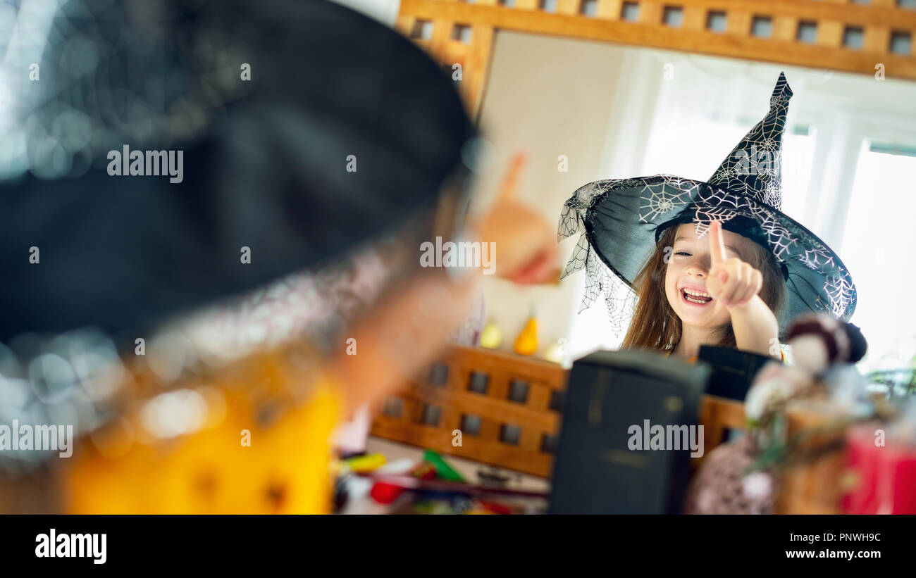 Adorable young girl wearing witch hat looking at herself in the mirror, pointing with finger and laughing. Happy child in halloween costume looking in Stock Photo
