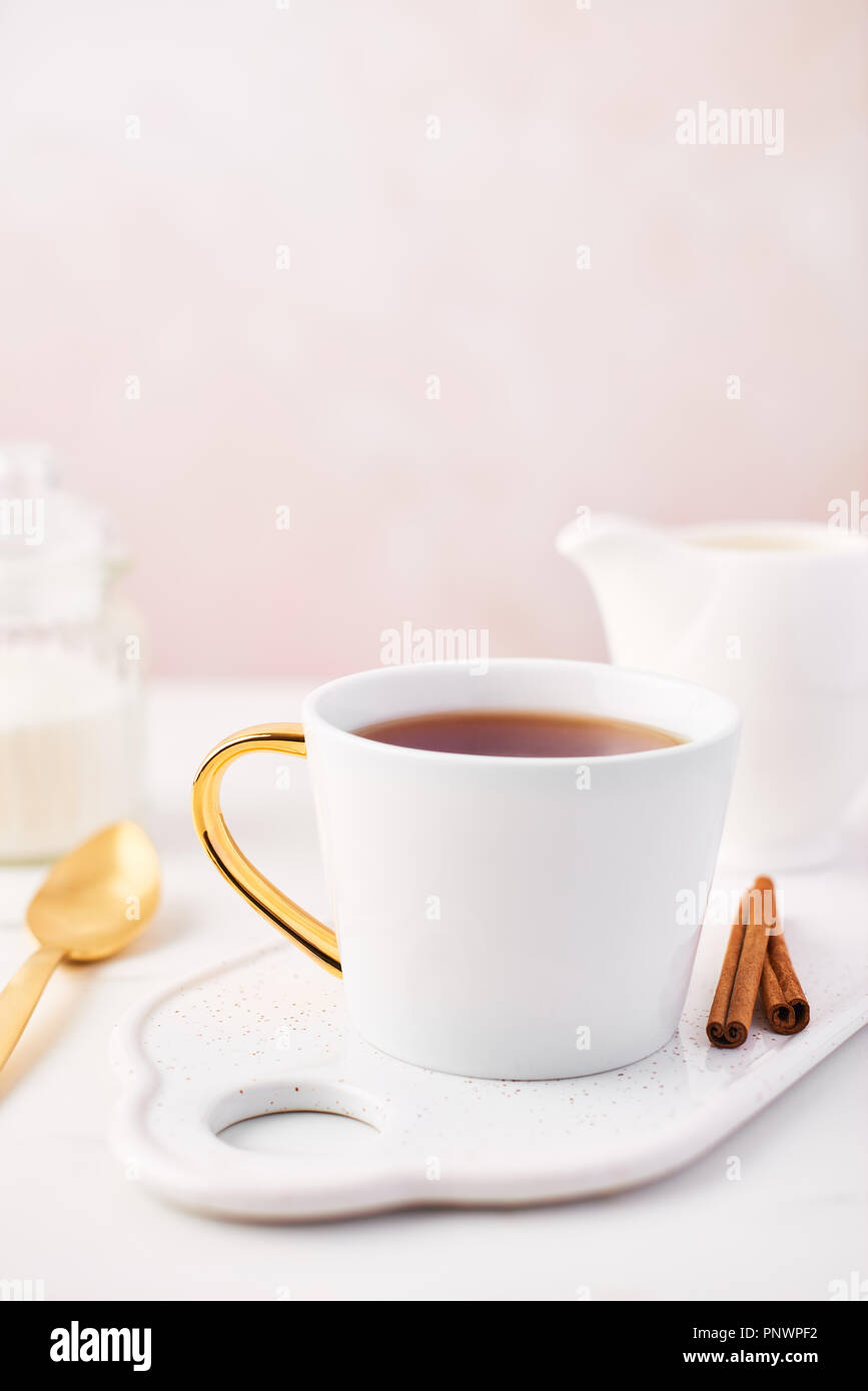 White cup of tea, cinnamon and milk jug on serving plate, golden spoon and sugar on white marble table top. Feminine rose background with copy space.  Stock Photo